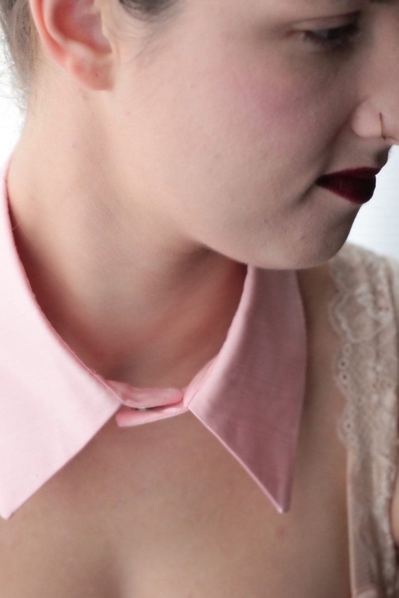 May include: A close-up of a person wearing a pink collared shirt with a button closure. The collar is large and has a pointed shape. The person is wearing a white lace top underneath the shirt.