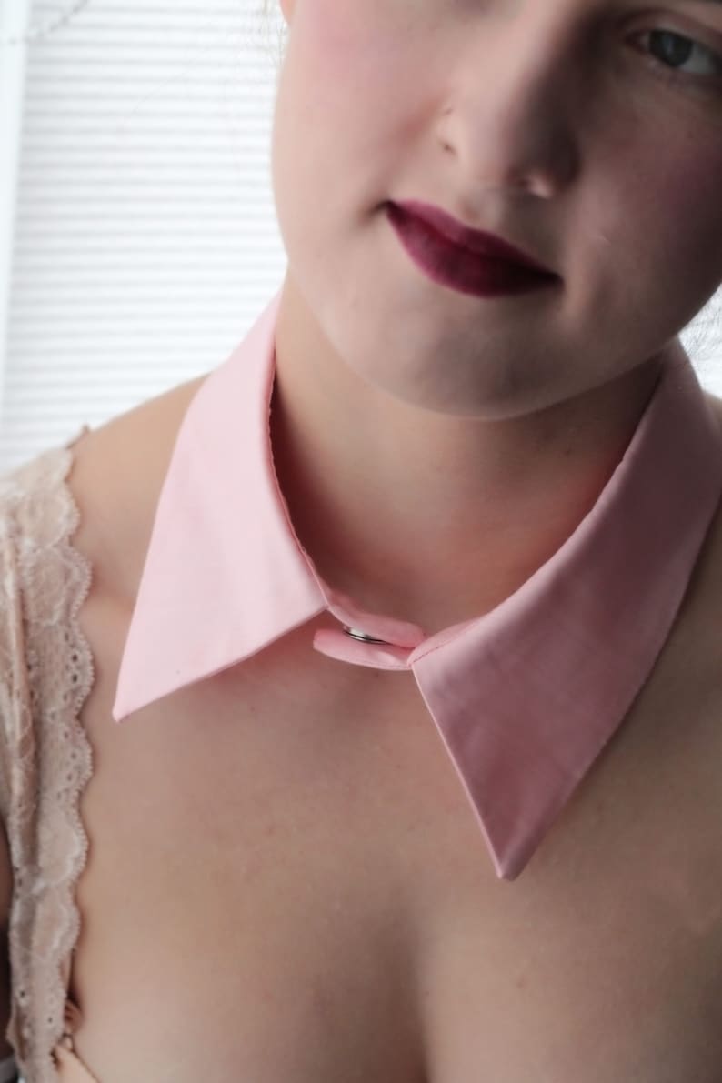 May include: A pink fabric collar with a silver button closure. The collar is worn over a white lace top.