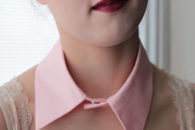 May include: A pink fabric collar with a button closure. The collar is worn over a white lace top.