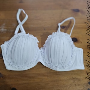 Op de afbeelding: Een witte beha met ruches en geplooide cups. De beha heeft verstelbare bandjes en ligt op een houten oppervlak. De tekst "choose just one or the set" staat aan de rechterkant.