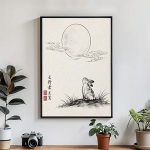 May include: Black framed art print featuring a rabbit gazing at a full moon. The artwork is a black and white ink drawing with Japanese text and red stamps. The rabbit sits on a small hill with grass, and clouds surround the moon.