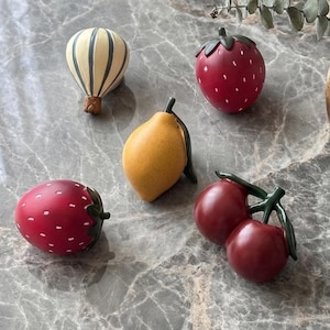 Könnte beinhalten: Eine Sammlung dekorativer Früchte und ein Heißluftballon-Figur sind auf einer Marmoroberfläche angeordnet. Die Früchte umfassen rote Erdbeeren mit weißen Punkten, eine gelbe Zitrone und rote Kirschen. Im Hintergrund steht eine Vase mit Trockenblumen.
