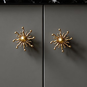 Könnte beinhalten: Zwei goldfarbene Sonnenaufgangs-Schrankknöpfe an einer grauen Schranktür. Die Knöpfe haben eine zentrale Kugel mit strahlenförmigen Strahlen. Die Schranktüren sind Teil eines größeren Schranksatzes mit einer schwarz-weißen Arbeitsplatte.