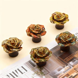 Pode incluir: Cinco puxadores de armário em cerâmica em forma de rosa em tons de castanho, laranja e verde. Cada puxador apresenta um design de rosa detalhado sobre uma base cilíndrica castanha escura. Os puxadores estão dispostos numa superfície clara.