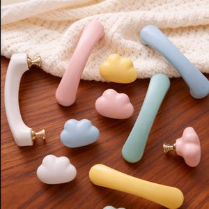 Puede incluir: Una colección de herrajes para armarios de colores pastel, que incluye tiradores y pomos en forma de nube. Los tiradores son de color rosa, azul, verde y amarillo, con herrajes dorados. Los pomos son de color rosa, amarillo y azul.