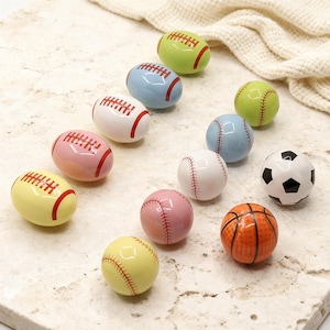 Op de afbeelding: Een verzameling keramische sportbalfiguren, waaronder American footballs, honkballen, een voetbal en een basketbal. De ballen zijn in verschillende kleuren zoals groen, blauw, roze, wit, geel en oranje. De figuren zijn gerangschikt op een lichtgekleurd oppervlak.