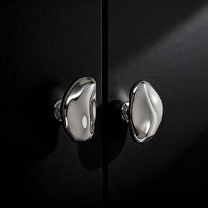 Puede incluir: Dos tiradores de armario cromados pulidos con forma orgánica de guijarro. Los tiradores están montados sobre una superficie oscura, posiblemente negra. Los tiradores tienen una superficie reflectante.