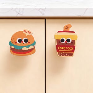 Könnte beinhalten: Zwei Schrankknöpfe im Cartoon-Stil. Einer ist ein Hamburger mit dem Wort "CUTE" oben. Der andere ist ein Soda-Becher mit den Worten "Coke cola". Beide Knöpfe haben große, runde Augen und sind vor einem hellbeigen Schrank angebracht.