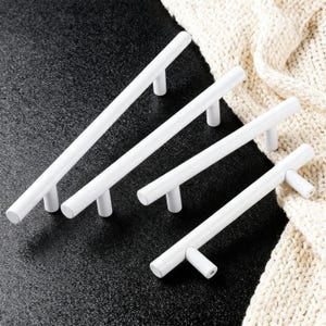 Könnte beinhalten: Vier weiße Schrankgriffe mit zylindrischer Stange und zwei Stützpfosten. Die Griffe sind diagonal auf einer schwarzen Oberfläche angeordnet, mit einem cremefarbenen Strickstoff im Hintergrund. Ideal für Küchen- oder Badezimmerschränke.