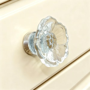 Peut inclure: Bouton de meuble en verre transparent en forme de fleur, avec une base argentée. Le bouton est monté sur une porte d'armoire crème. La fleur a des pétales détaillés et un centre texturé, ajoutant une touche décorative.