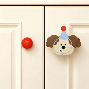 May include: Two cabinet knobs. One is a solid, glossy red sphere. The other is a decorative, cartoon-style dog face with brown ears, a blue party hat, and a blue bow tie. Both are mounted on a white cabinet door.