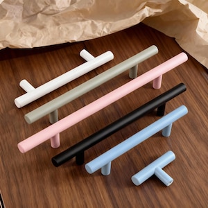 Könnte beinhalten: Eine Auswahl an farbigen Schrankgriffen in T-Form, in Weiß, Grau, Rosa, Schwarz und Blau. Die Griffe sind auf einer Holzoberfläche angeordnet, um ihre verschiedenen Farben und Designs zu präsentieren. Maße in Zentimetern.