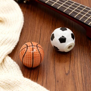 Op de afbeelding: Twee decoratieve kastknoppen. Eén is een basketbalontwerp met oranje en zwarte details. De andere is een voetbalontwerp met zwarte en witte details. Beide liggen op een houten oppervlak.