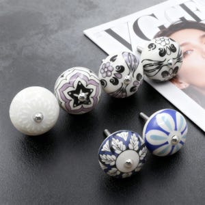 Peut inclure: Ensemble de boutons de meubles décoratifs en céramique. Les boutons présentent divers motifs floraux et géométriques en blanc, violet et bleu, avec des garnitures argentées. Les boutons sont disposés sur une surface sombre.