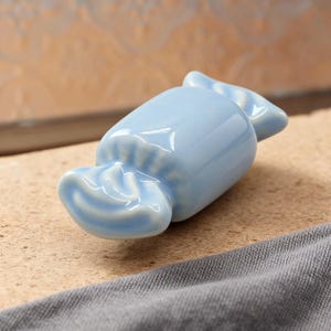 Könnte beinhalten: Ein hellblaues, keramikartiges, bonbonförmiges Dekorationsobjekt. Das glänzende Bonbon liegt auf einer strukturierten Oberfläche. Die Enden des Bonbons sind gefaltet, um einem verpackten Bonbon zu ähneln.