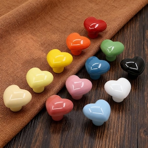May include: A collection of heart-shaped cabinet knobs in various colors, including red, orange, yellow, green, blue, black, pink, and white. The knobs are arranged on a brown surface, showcasing their glossy finish and decorative design.