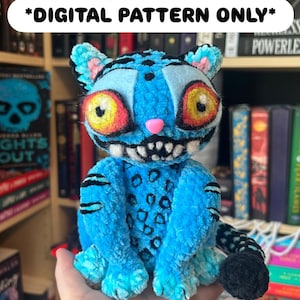 May include: A handmade blue plush cat with black stripes and a pink nose. The cat has large yellow and red eyes and a toothy grin. The text "*DIGITAL PATTERN ONLY*" is at the top. The cat is sitting in front of a bookshelf.