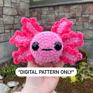 Pode incluir: Brinquedo de pelúcia axolotl de crochê rosa com grandes olhos pretos. O brinquedo está sendo segurado por uma mão. O texto "*DIGITAL PATTERN ONLY*" é visível abaixo do brinquedo.
