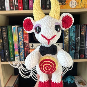 May include: White crocheted goat amigurumi with yellow horns, red ears, black bow tie, and a red and yellow swirl design on its belly.