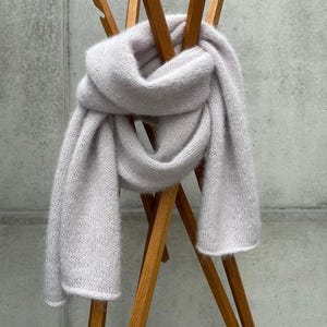 Puede incluir: Una bufanda de punto gris claro drapeada sobre un perchero de madera. La bufanda está hecha de un material suave y esponjoso y está anudada en el medio. El perchero tiene un diseño simple y minimalista con múltiples brazos de madera.