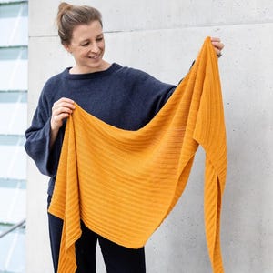 May include: Mustard yellow knitted shawl with a ribbed texture. The shawl is held up, displaying its triangular shape and the detailed knit pattern. The color is a warm, inviting shade, suitable for fall or winter wear. The shawl is a versatile accessory.