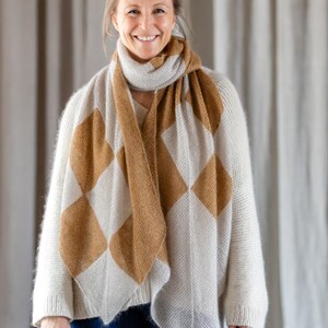Könnte beinhalten: Eine Frau trägt einen cremefarbenen Pullover und Schal mit einem braunen und cremefarbenen Rautenmuster. Der Schal ist um den Hals und über den Schultern drapiert. Die Frau lächelt und trägt eine blaue Jeans.