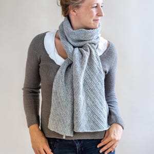 Könnte beinhalten: Ein hellgrauer Strickschal mit einem strukturierten, diagonalen Muster. Der Schal ist um den Hals und über die Schultern drapiert. Die Person trägt einen grauen Pullover und blaue Jeans.