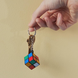 May include: A keychain with a miniature Rubik's Cube attached. The cube features the classic color scheme of red, blue, green, orange, yellow, and white. The keys are made of brass and are attached to a silver key ring.