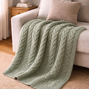 Puede incluir: Una manta de punto trenzado verde salvia sobre un sofá de color claro. La manta presenta un diseño tejido texturizado. Dos cojines de tonos neutros descansan sobre el sofá. La manta está sobre una alfombra beige.