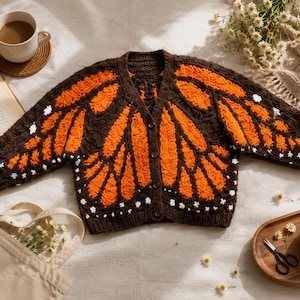 May include: A brown knitted cardigan with orange butterfly wing designs. The sweater has a button-up front and white accents. The image also includes a cup of coffee, a book, and a tote bag.