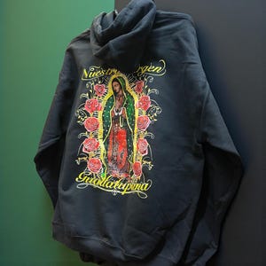 May include: Black hoodie with a graphic of Our Lady of Guadalupe and roses. The text "Nuestra Virgen Guadalupana" is in gold. The hoodie has a hood and a relaxed fit. The design is on the back of the garment.