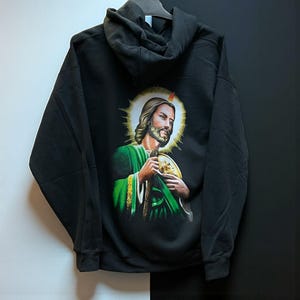 May include: Black hooded sweatshirt with a religious image on the back. The graphic shows a man with a halo, wearing green and gold robes. The sweatshirt has a drawstring hood and long sleeves. The garment is hanging on a hanger.