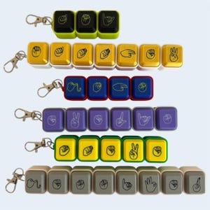 May include: Six rows of keycap-style keychains featuring American Sign Language hand signs printed on the surface. The rows are arranged in different colors including black, yellow, blue, purple, green, and gray, each with a metal lobster clasp attached.