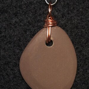 May include: A brown, smooth, pebble-shaped pendant with a small hole. The pendant is wrapped with copper wire and attached to a silver-colored bail. The background is a dark, textured surface.