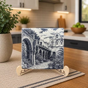 May include: A square, blue and white tile featuring a detailed architectural illustration of a building with arches, a bell tower, and lush greenery. The tile is displayed on a decorative wooden stand with a curved design.