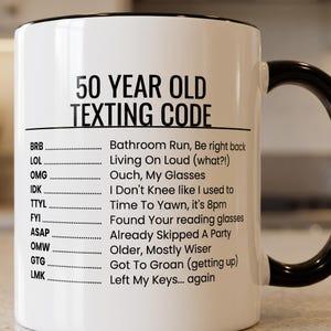 May include: A white ceramic mug with a black handle and rim, featuring the black printed text 50 YEAR OLD TEXTING CODE followed by a list of acronyms and humorous definitions including BRB, LOL, OMG, IDK, TTYL, FYI, ASAP, OMW, GTG, and LMK.