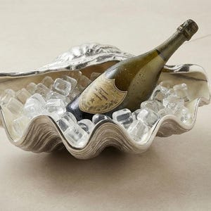 Könnte beinhalten: Eine große, silberfarbene Muschelschale, gefüllt mit klaren Eiswürfeln und einer einzelnen Glasflasche Dom Pérignon Champagner. Die Schale hat eine texturierte, geriffelte Außenseite und eine glatte, reflektierende Innenseite.