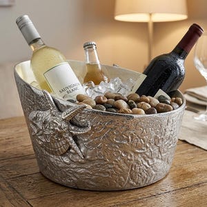 Op de afbeelding: Een zilveren wijnkoeler met een textuur, gevuld met ijs, rivierstenen en drie flessen wijn. Eén fles is witte wijn, een andere is rode wijn en de derde is een kleine fles bier. Een wijnglas en een kleine vetplant staan in de buurt.