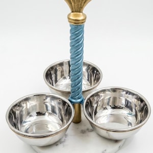 Puede incluir: Un soporte para servir de tres cuencos con una base circular de mármol blanco, tres cuencos de acero inoxidable pulido y un asa central vertical con una columna trenzada de color azul claro rematada por un adorno dorado en forma de concha.