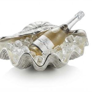 Könnte beinhalten: Eine silberfarbene, muschelförmige Metallschale, gefüllt mit klaren Eiswürfeln und einer Flasche Chateau Pontet-Canet Wein. Die Schale hat eine texturierte, gerippte Außenseite und eine glatte, reflektierende Innenfläche, die die Flasche hält.