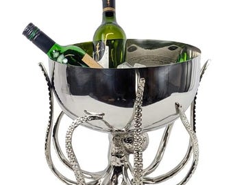 Puede incluir: Una cubitera de metal plateado pulido con forma de pulpo, con ocho tentáculos texturizados que forman la base y los lados. El cuenco contiene dos botellas de vino de cristal con etiquetas oscuras y cubitos de hielo.