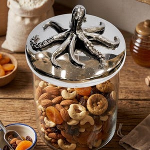 May include: A clear glass jar filled with nuts and dried fruit, topped with a silver octopus lid. A small tag reads "Reserve Blend - Dry Fruits". The jar sits on a wooden surface, with other food items in the background.