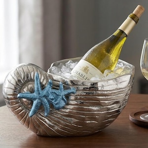 Op de afbeelding: Een zilverkleurige metalen wijnkoeler in de vorm van een nautilusschelp, met een getextureerd oppervlak en een decoratief blauw zeesteraccent. De koeler is gevuld met ijs en bevat een fles Chablis-wijn, vergezeld door twee wijnglazen.