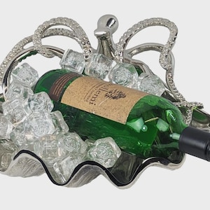 Op de afbeelding: Een zilverkleurige metalen wijnflessenhouder in de vorm van een schelp met een sierlijk, getextureerd handvat. Een groene glazen wijnfles met een beige etiket met de tekst 'Tollemi' ligt in de houder, omringd door heldere, gefacetteerde acryl ijsblokjes.