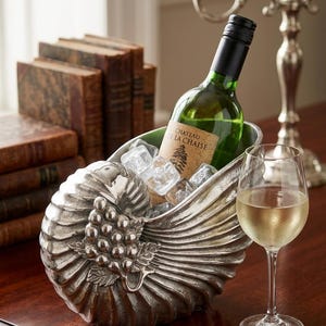 Op de afbeelding: Een zilverkleurige wijnkoeler in de vorm van een nautilusschelp met een reliëf van een druiventros. De koeler bevat een groene glazen fles met het etiket Chateau de la Chaise en ijsblokjes. Een wijnglas en een druifvormige flesopener staan ernaast.
