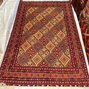 以下が含まれることがあります： 赤、金、茶色の色合いで繰り返されるダイヤモンド模様の長方形の手織りラグ。 ラグには、花柄と幾何学模様の細かいボーダーがあります。 ラグは白い表面に表示されています。