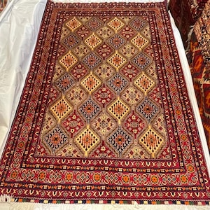 以下が含まれることがあります： 赤、金、茶色の色合いで繰り返されるダイヤモンド模様の手織りの長方形のラグ。 ラグには、花柄と幾何学模様の細かいボーダーがあります。 フリンジは白です。 ラグはおそらくウール製です。