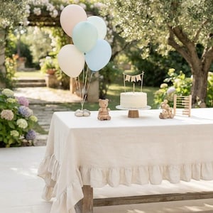 Op de afbeelding: Een beige linnen tafelkleed met een geplooide rand over een houten tafel. Op de tafel staan een tros pastelkleurige ballonnen, een houten berenfiguurtje, een witte taart met een banner met de tekst ONE, een houten speelgoedtrein en een kleine telraam.