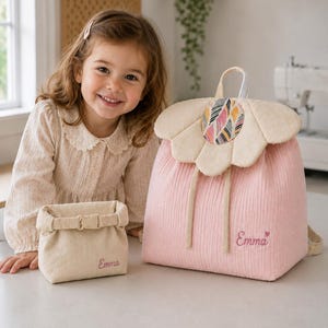 May include: A light pink backpack with a large beige flower-shaped flap featuring a colourful patterned centre, and a matching beige roll-top pouch. Both items are embroidered with the name Emma in pink script, with a small heart icon.
