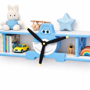 Puede incluir: Estantería azul y blanca con forma de avión con hélice, que contiene juguetes y libros. La estantería tiene dos niveles y está decorada con una cara sonriente. Los juguetes incluyen un conejito de peluche, un coche amarillo y un cohete de madera.